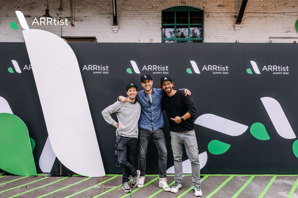 Drei Personen posieren vor einer Wand mit dem Logo der ARRtist Summit 2023.